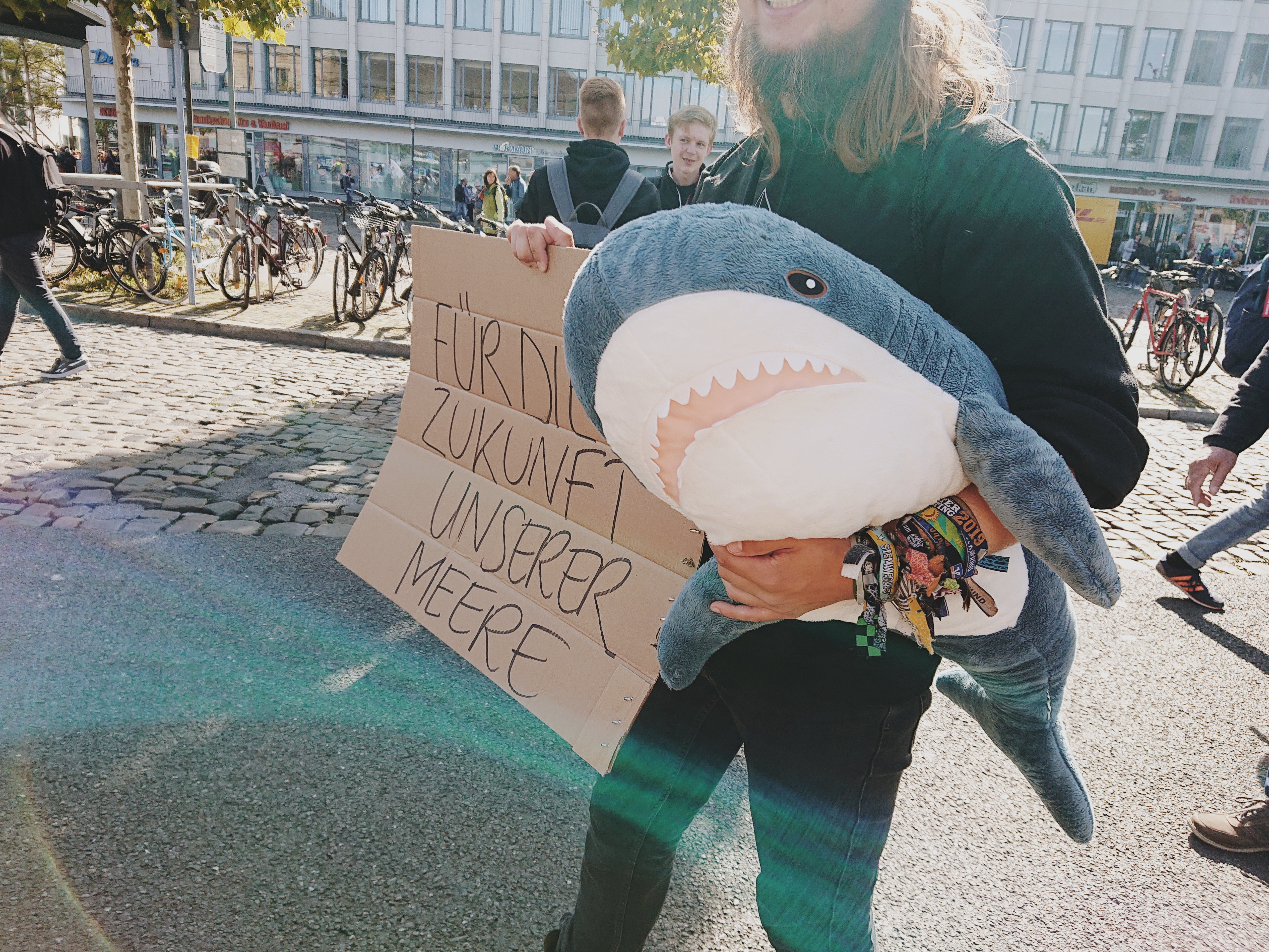 Eine Person steht auf einem öffentlichen Platz. In der Hand hält sie einen Stofftier-Hai und ein selbstgemaltes Plakat mit der Aufschrift "Für die Zukunft unserer Meere"..